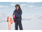 Kind in ski-jas, skibroek, helm en handschoenen, past een handschoen aan met skistokken in de buurt.