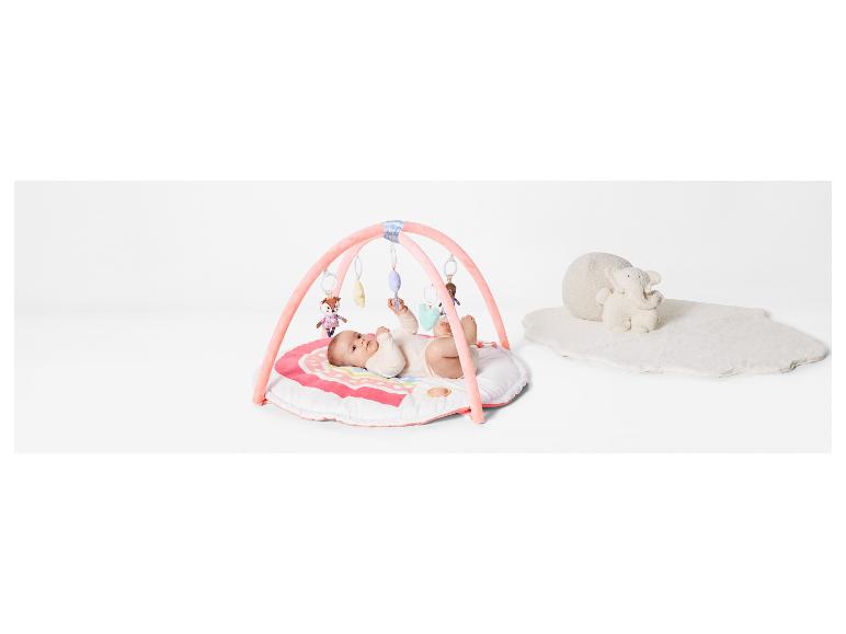 Een baby ligt op een roze speelmat met een boog met speelgoed en een zachte witte mat met een olifant.