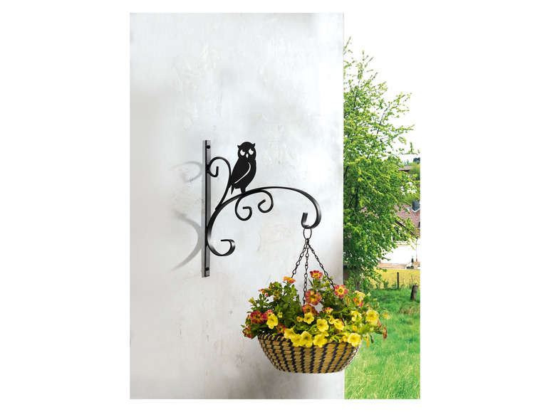 Een hangende plantenbak met bloemen aan een metalen haak met een uilvorm.