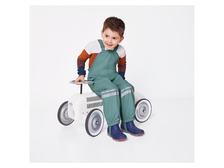 Een kind in een groene overall en blauwe laarzen zit op een wit speelgoedautootje.