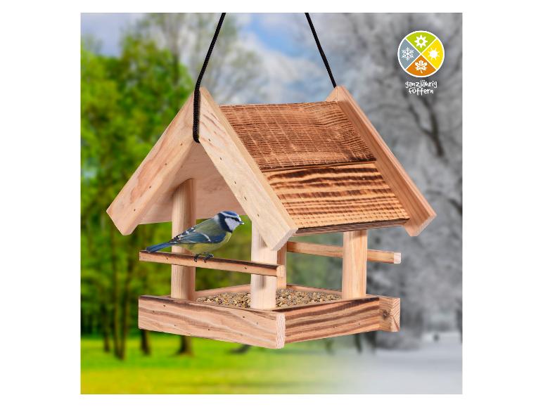 Houten vogelhuisje met een pimpelmees die zaden eet, tegen een zomer- en winterachtergrond.