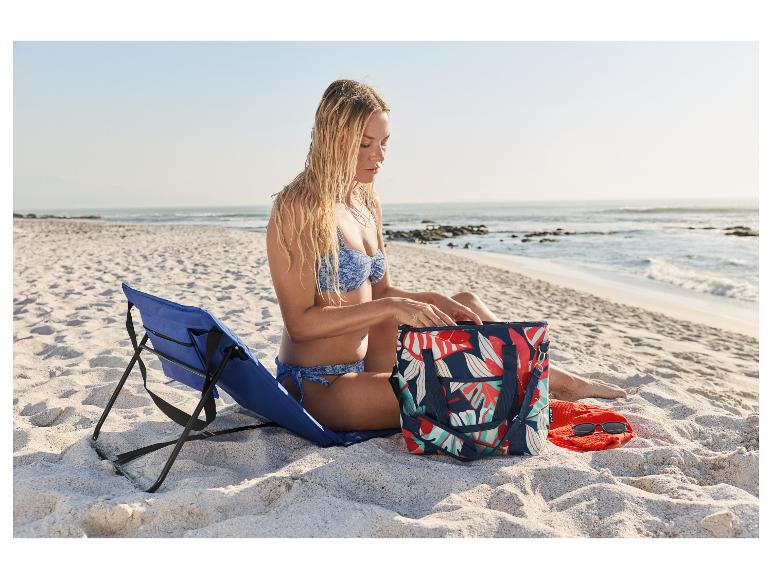Vrouw in bikini op strand, zittend naast een blauwe strandstoel en een tas met tropische print.
