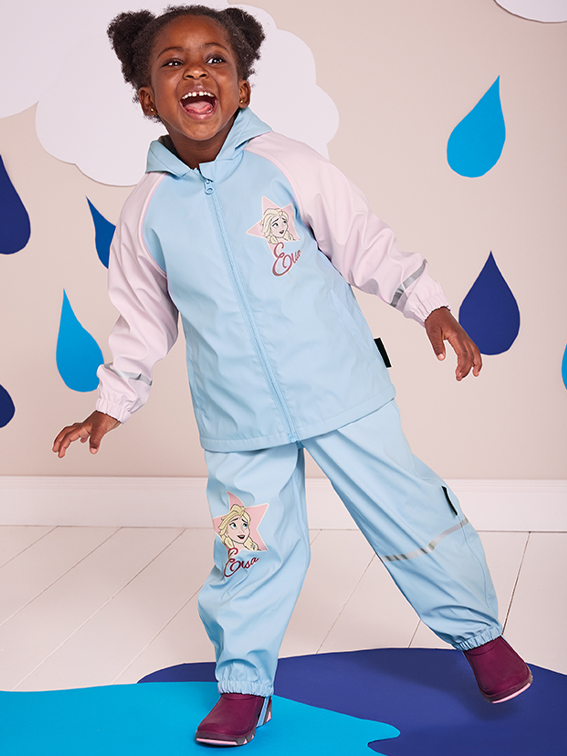 Een kind in een lichtblauw en roze Frozen-regenpak, lachend en staand op een blauwe mat.