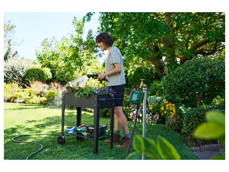 Vrouw besproeit planten in een verhoogde moestuinbak met een slang, naast een besproeiingstimer.