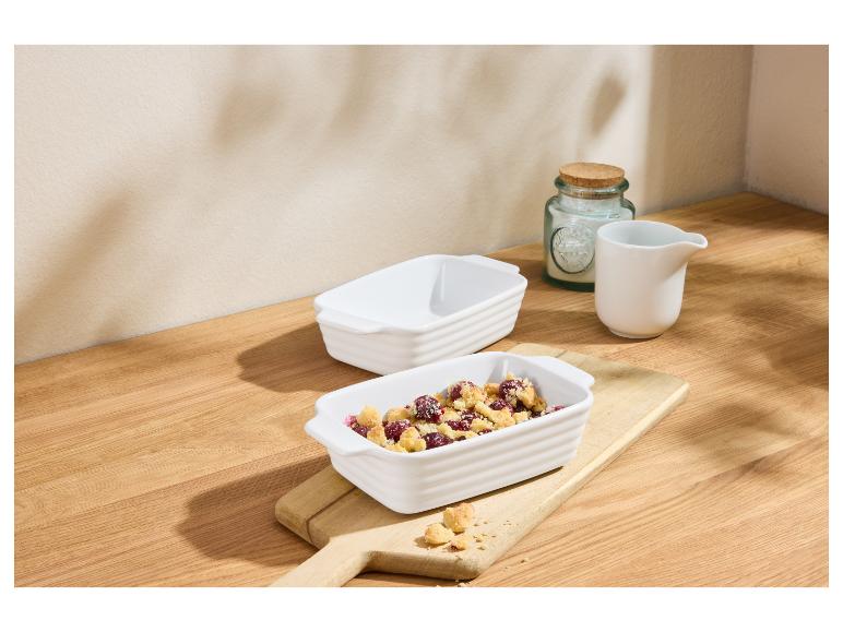 Twee witte ovenschaaltjes, één met bessen-crumble dessert, op een houten plank.