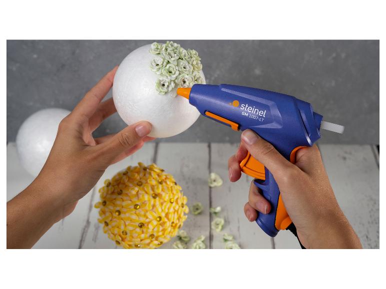 Lijmpistool bevestigt witte bloemen op een piepschuimbal, een andere bal is versierd met gele bloemen.