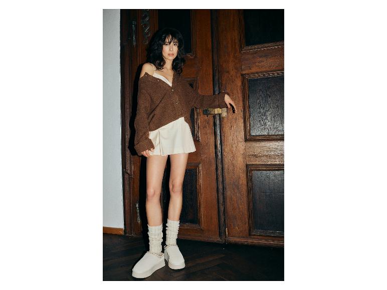 Vrouw in bruin vest, witte top en geplooide minirok, met witte pantoffels en sokken.