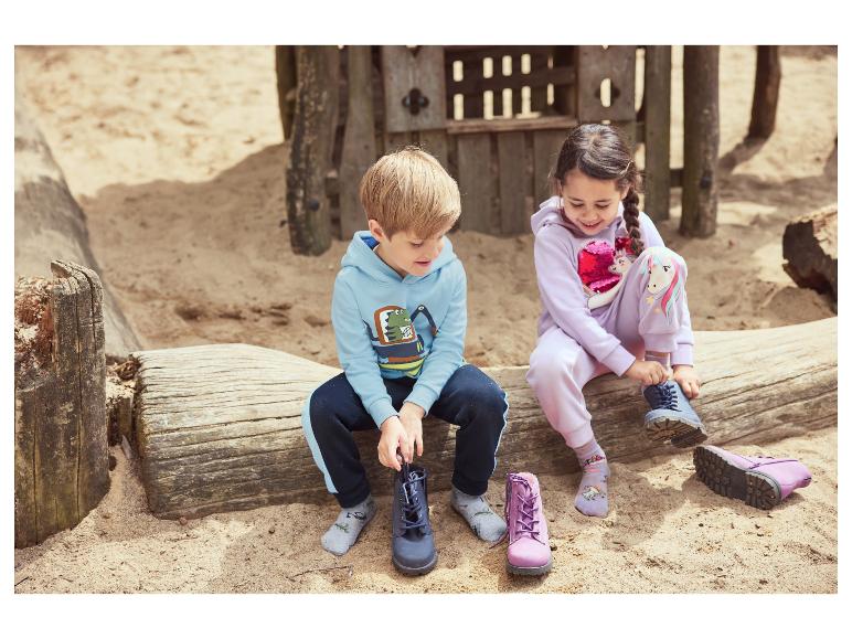 Twee kinderen trekken laarzen aan op een houten boomstam op een zandige speeltuin.