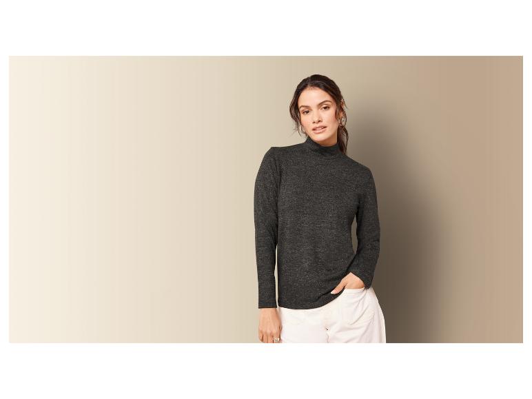 Een vrouw draagt een donkergrijze trui met hoge hals en witte broek.