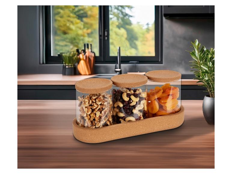 Drie glazen potten met kurken deksels gevuld met walnoten, mix noten en gedroogde abrikozen, op een kurken dienblad.