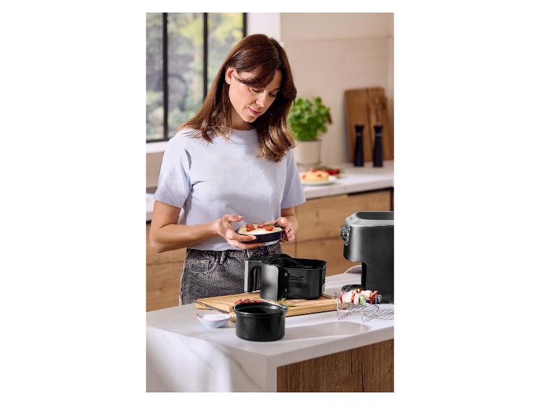 Vrouw bereidt maaltijd in keuken, houdt een kleine ovenschaal vast, met een airfryer ernaast.