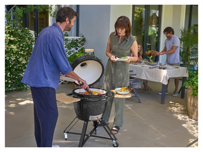 Mensen grillen voedsel op een keramische barbecue op een terras, met een eettafel op de achtergrond.