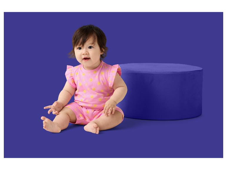Een baby in een roze romper met zonnepatroon zit naast een blauwe poef.