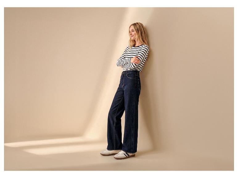 Vrouw in gestreept shirt en wijde jeans leunt tegen een muur, met sneakers aan.