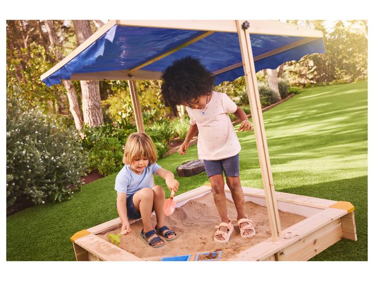 Twee kinderen spelen in een houten zandbak met blauwe luifel in een tuin.