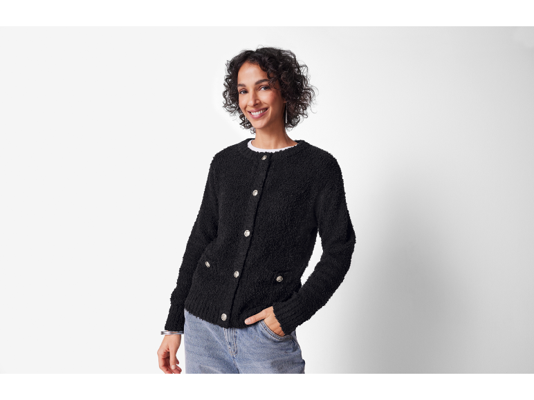 Lachende vrouw in een zwart getextureerd vest en jeans, hand in zak.