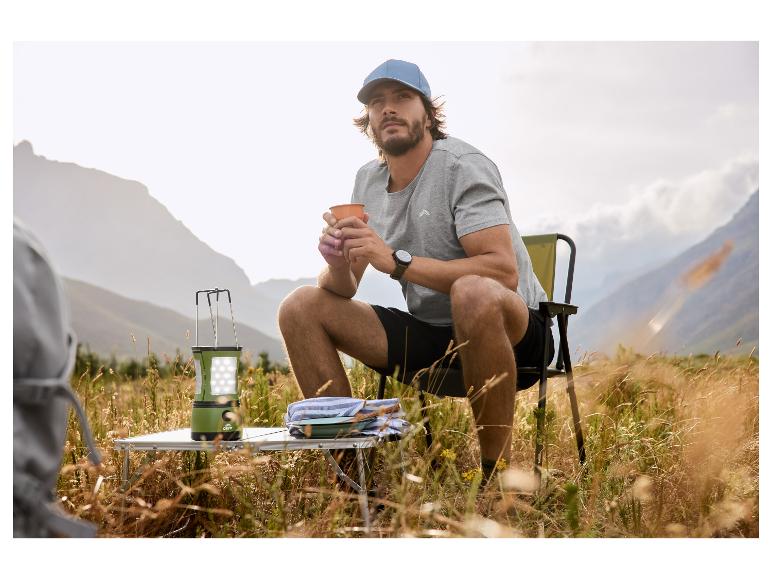 Man in pet en T-shirt zit op campingstoel met mok, naast een lantaarn en tafel.