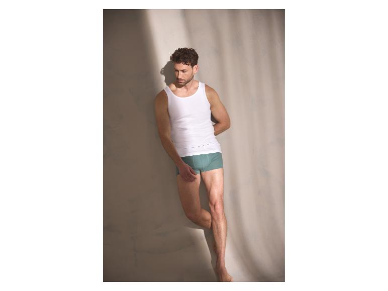 Man die tegen een muur leunt, gekleed in een herenboxershort en een hemd.