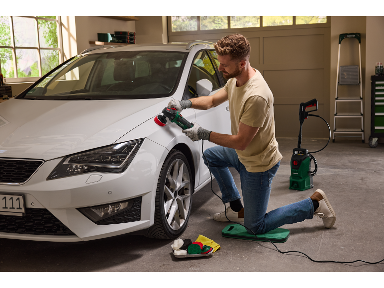 Een man polijst een witte auto met een roterende polijstmachine in een garage.