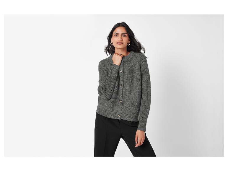 Een vrouw draagt een grijze gebreide trui met knopen en een zwarte broek.