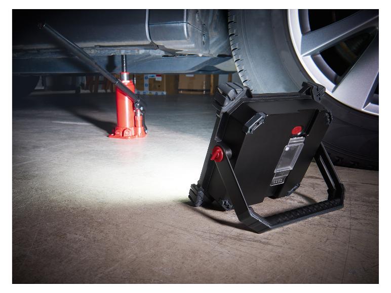 Een rode krik tilt een auto op, verlicht door een zwarte werklamp op de vloer.