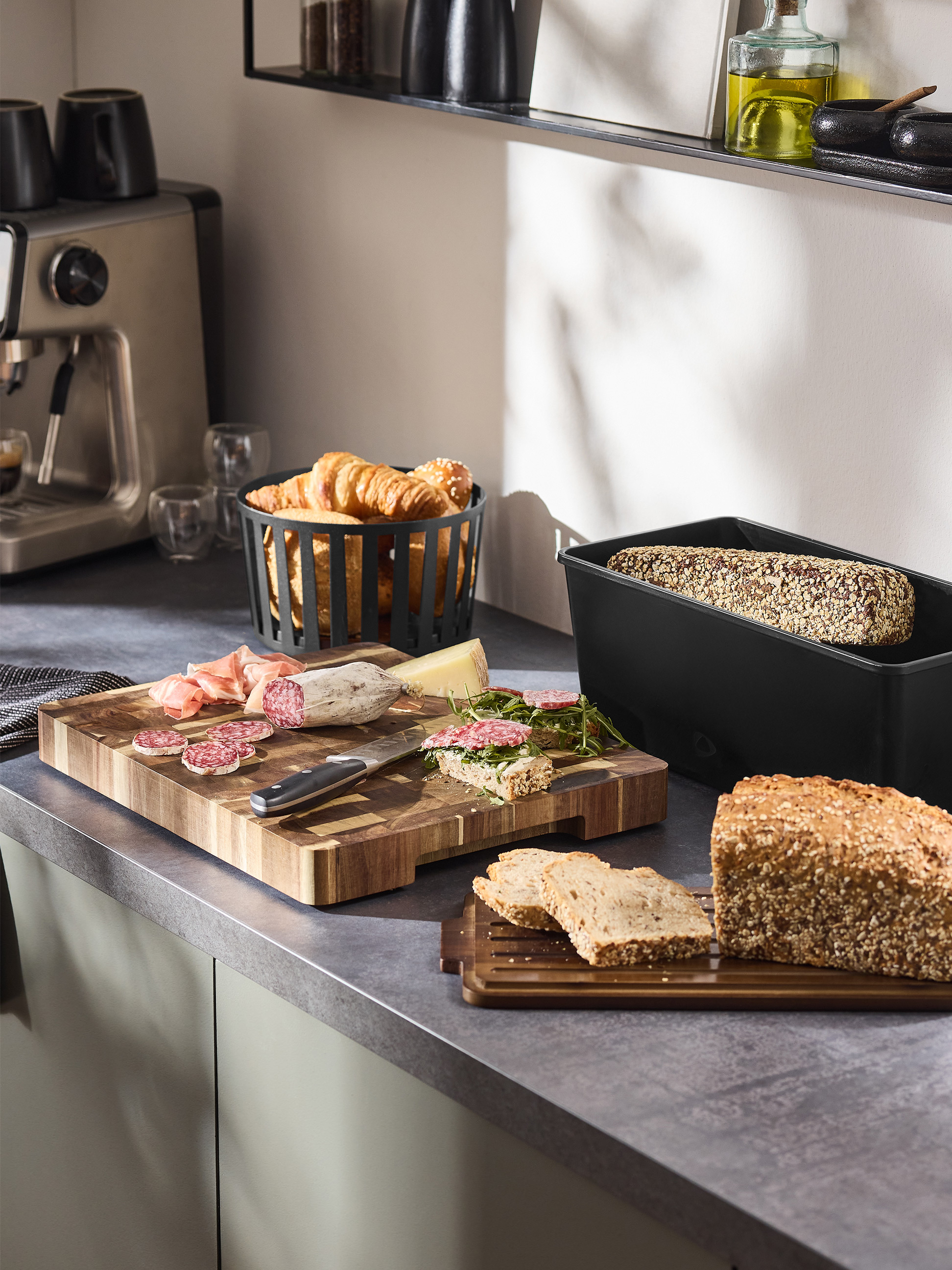Ontbijt met koffiezetapparaat, brood, vleeswaren en croissants op een aanrecht.