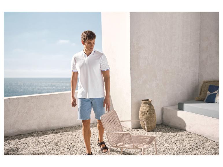 Man in wit poloshirt en spijkerbroek op een terras met uitzicht op zee.