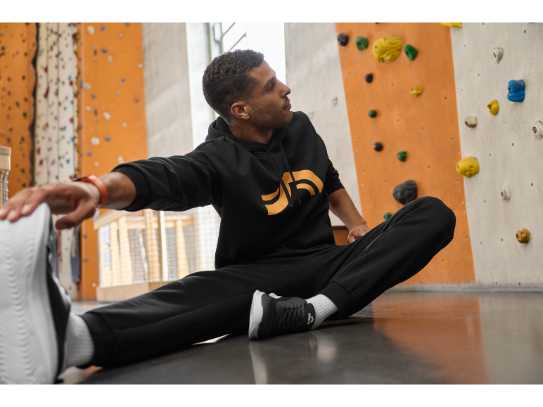 Man in zwarte hoodie en joggingbroek rekt zich uit op de vloer voor een klimmuur.