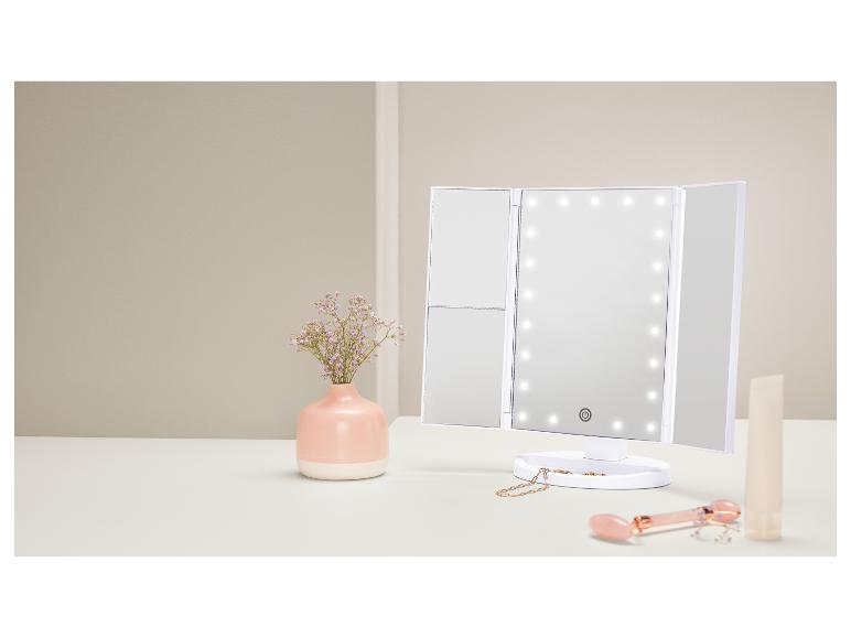 Driedelige make-upspiegel met LED-verlichting, vaas met bloemen en schoonheidsproducten op tafel.