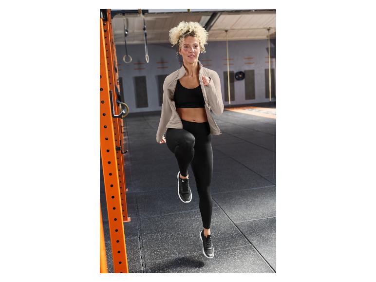 Vrouw in zwarte sportkleding en beige jas, trainend in een sportschool.