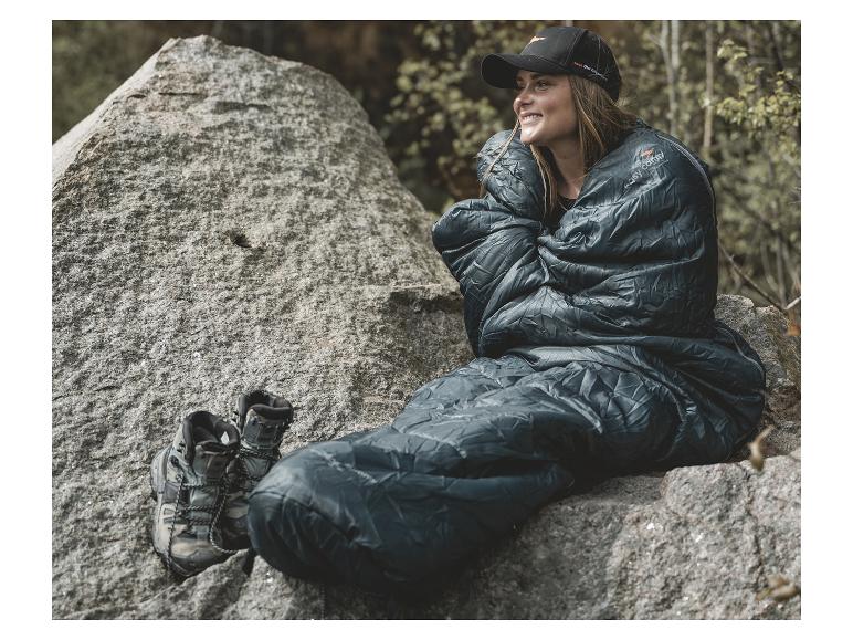 Een vrouw die op een rots zit, omhuld door een Easy Camp slaapzak.