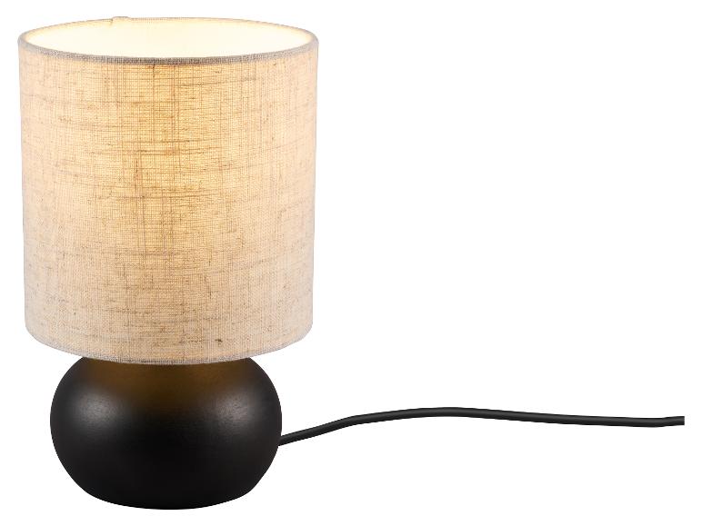 Tafellamp met een zwarte bolvormige voet en een cilindrische beige stoffen kap, verlicht.
