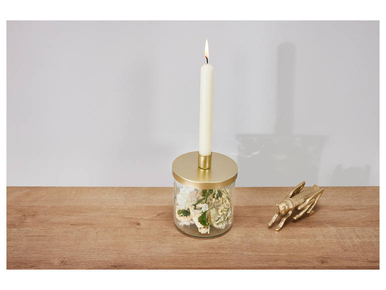 Een brandende kaars in een glazen pot met gedroogde bloemen en een gouden sprinkhaan op een houten tafel.