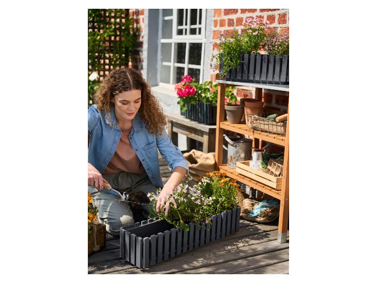 Vrouw plant bloemen in een grijze plantenbak op een houten terras, met een tuinrek.
