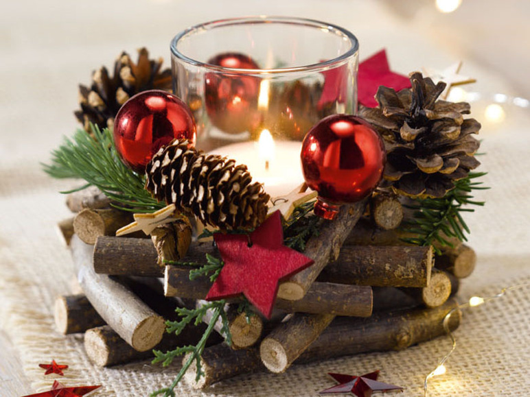 Een kerstkaars in een glazen houder, versierd met dennenappels, takken en rode kerstballen.