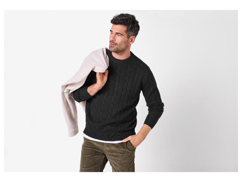 Man in een zwarte kabeltrui, corduroy broek en een beige jas over zijn schouder.