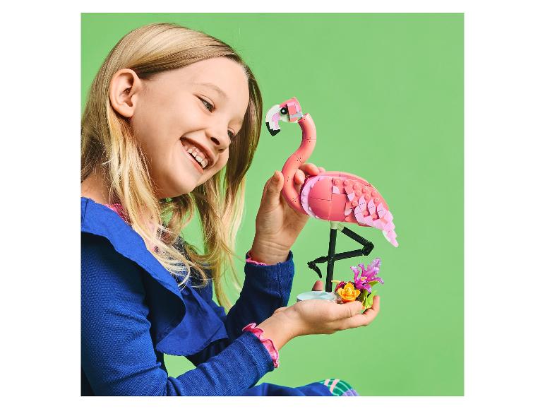 Een lachend meisje houdt een roze bouwsteenflamingo en bloemen vast.