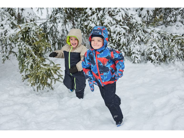 Twee jongens in winteroveralls en jassen rennen in de sneeuw naast dennenbomen.
