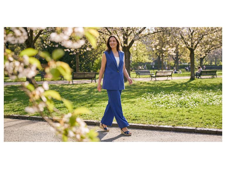Vrouw in blauw pak met gilet en wijde broek loopt door een park.