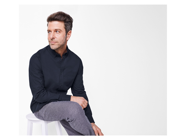 Een man in een donkerblauw overhemd en grijze broek zit op een witte kruk.