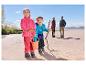 Twee kinderen in fleece jassen en outdoor broeken spelen op een zandstrand, ouders op de achtergrond.