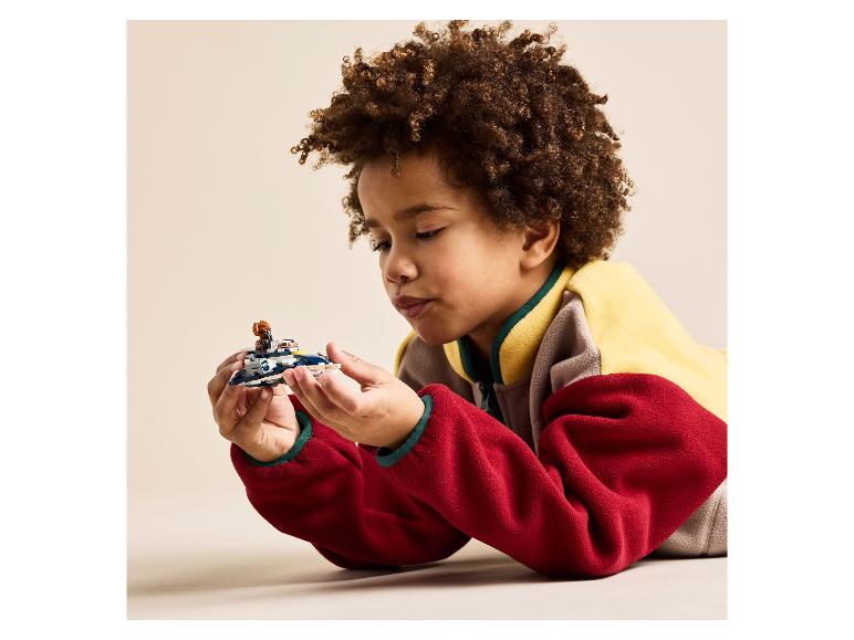 Een kind met krullend haar, liggend op de buik, speelt met een Lego ruimteschip.