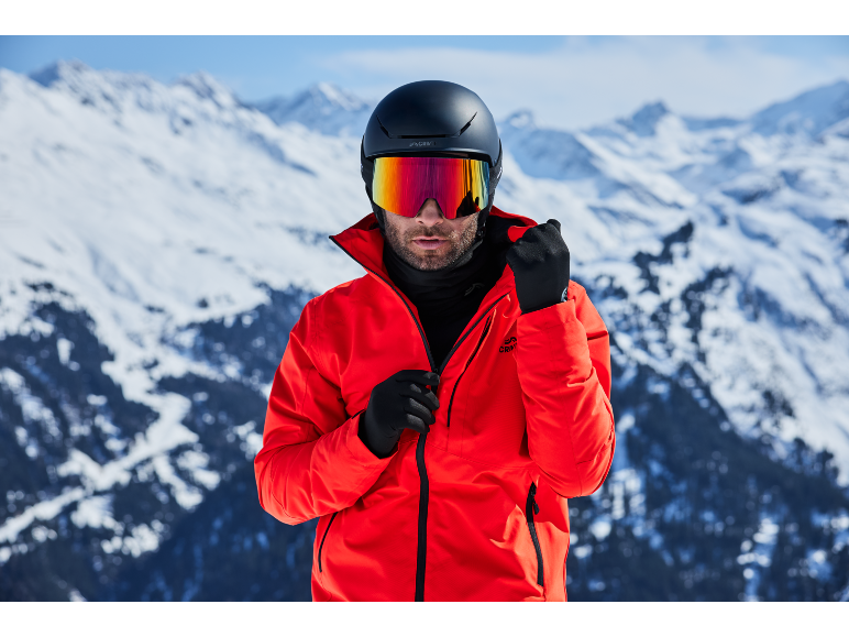 Man met skihelm, skibril en rode jas in besneeuwde bergen.