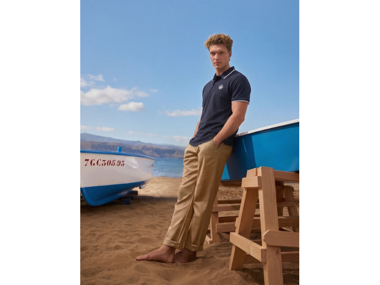 Man in marineblauwe polo en beige broek op het strand met boten.