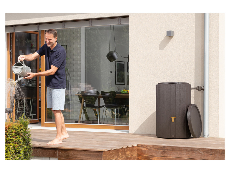 Een man giet water uit een gieter op een plant, naast een regenton