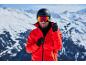 Man met skihelm, skibril en rode jas in besneeuwde bergen.