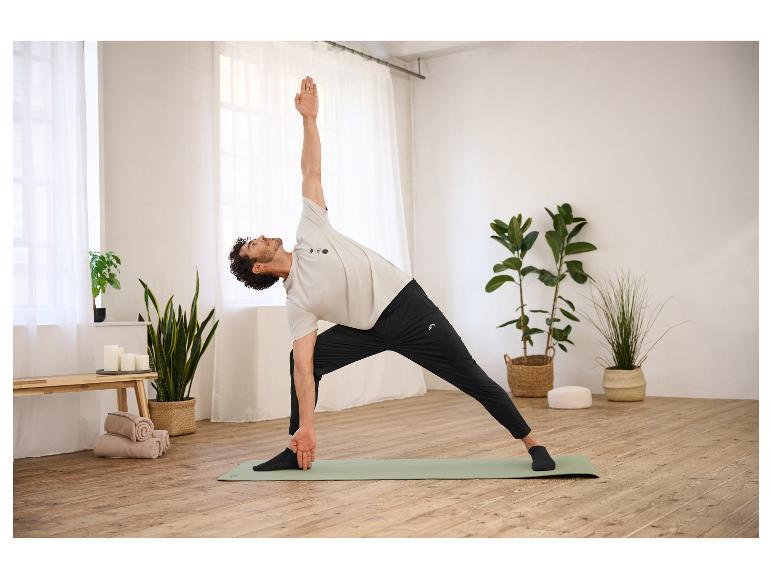 Een man in een CRIVIT functioneel shirt doet yoga op een mat in een lichte kamer.