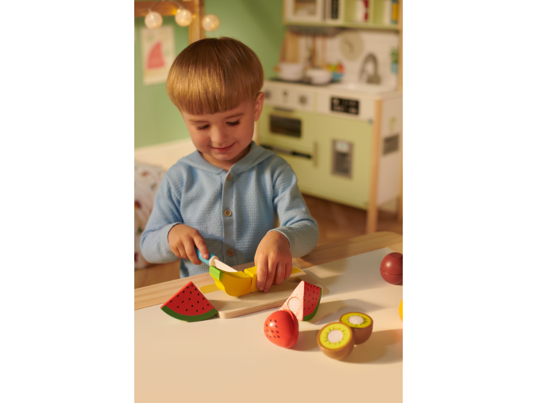 Peuter speelt met speelgoedfruit en snijplank in een speelse keukenomgeving.