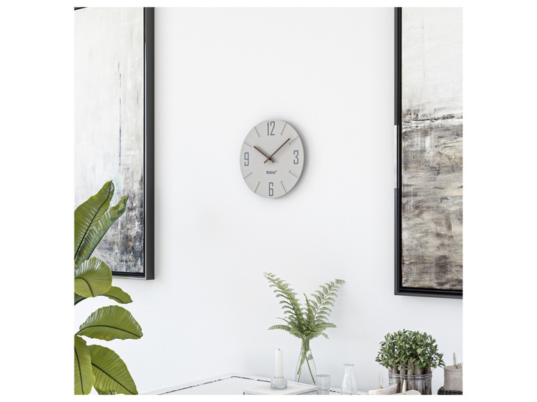 Een witte Mebus wandklok met twee schilderijen ernaast.