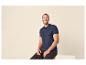 Lachende man in marineblauwe poloshirt en donkere jeans, zittend op een lichte ondergrond.